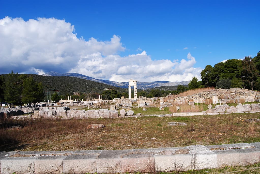 A landscape image featuring ancient greek ruins. In the distance some tall structures can be seen. Behind the ruins are mountains. The image features a stunning blue sky with low white clouds.