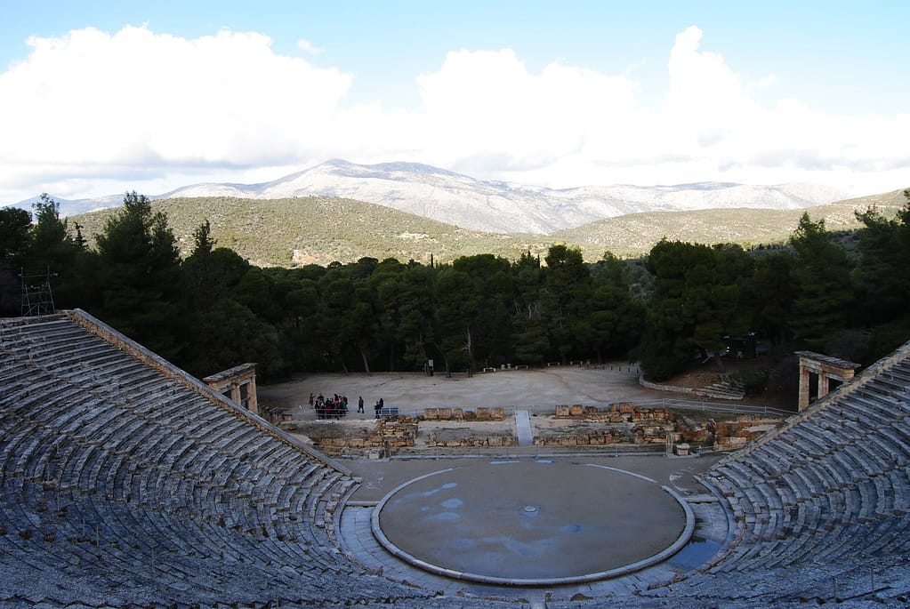 The view from the top of a tall ancient amphitheater. Snow capped mountains peer over trees in the distance.